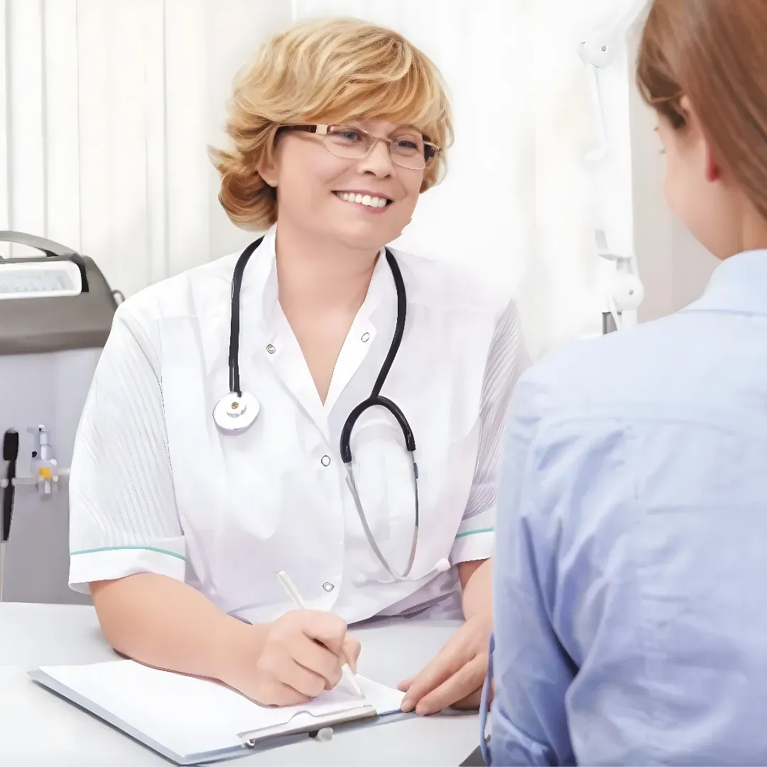 A female doctor sitting at a desk talking to a patient. The doctor is wearing a stethoscope around her neck and is holding a pen in her hand. On the desk in front of them is a book and a pen, and in the background there is a machine and a white curtain.
