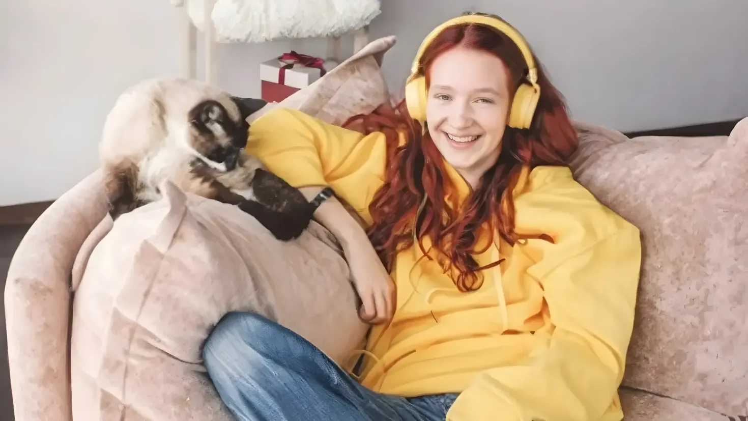 A woman sitting on a couch with a smile on her face, wearing headphones and holding a cat in her arms. Behind her is a wall with a gift box and other objects on a stand.