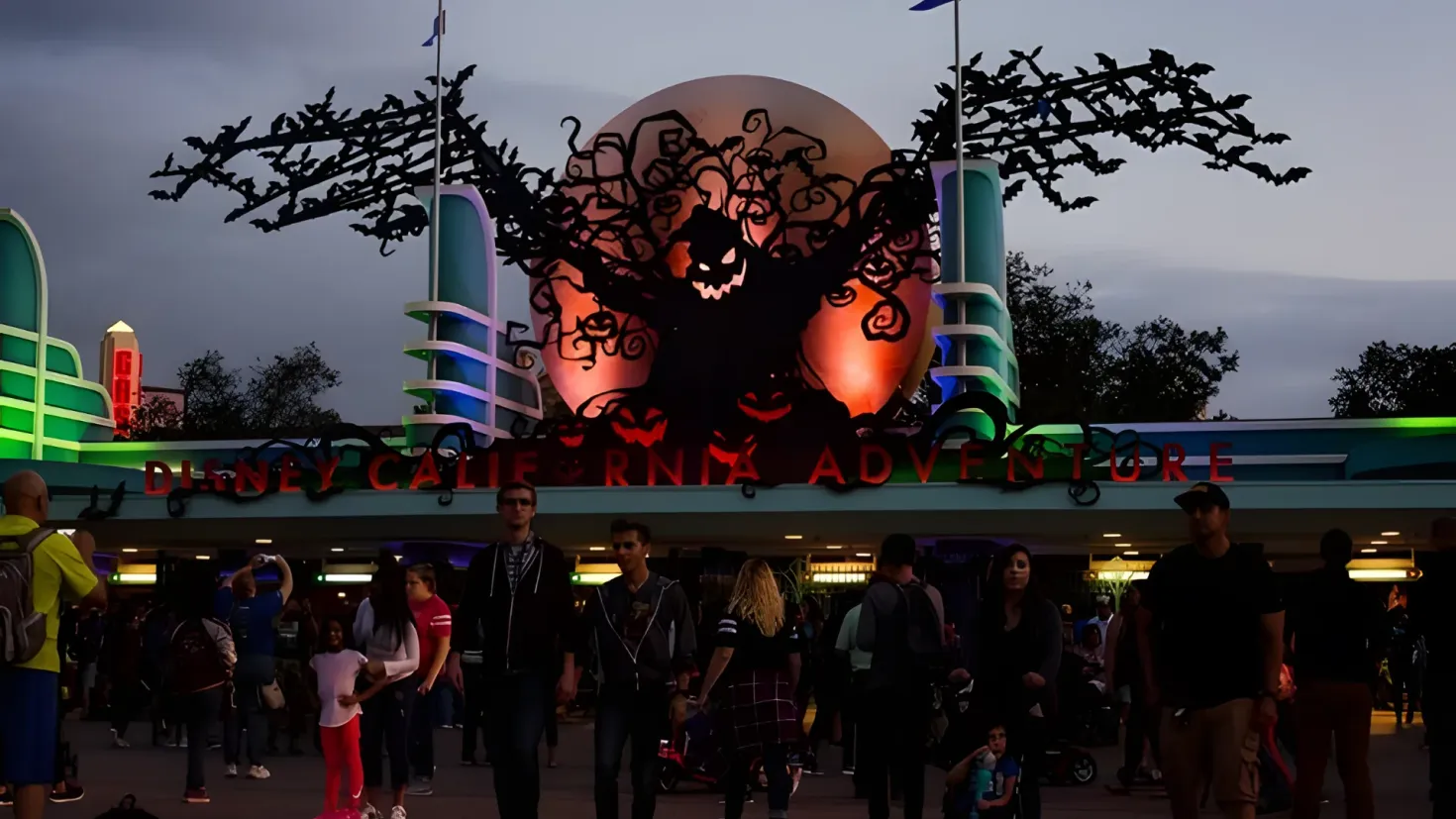 A group of people standing in front of a building at night. Some of them are wearing backpacks and hats, and there is a stroller in the foreground. The building has text on it, and in the background there are trees and a sky with clouds.