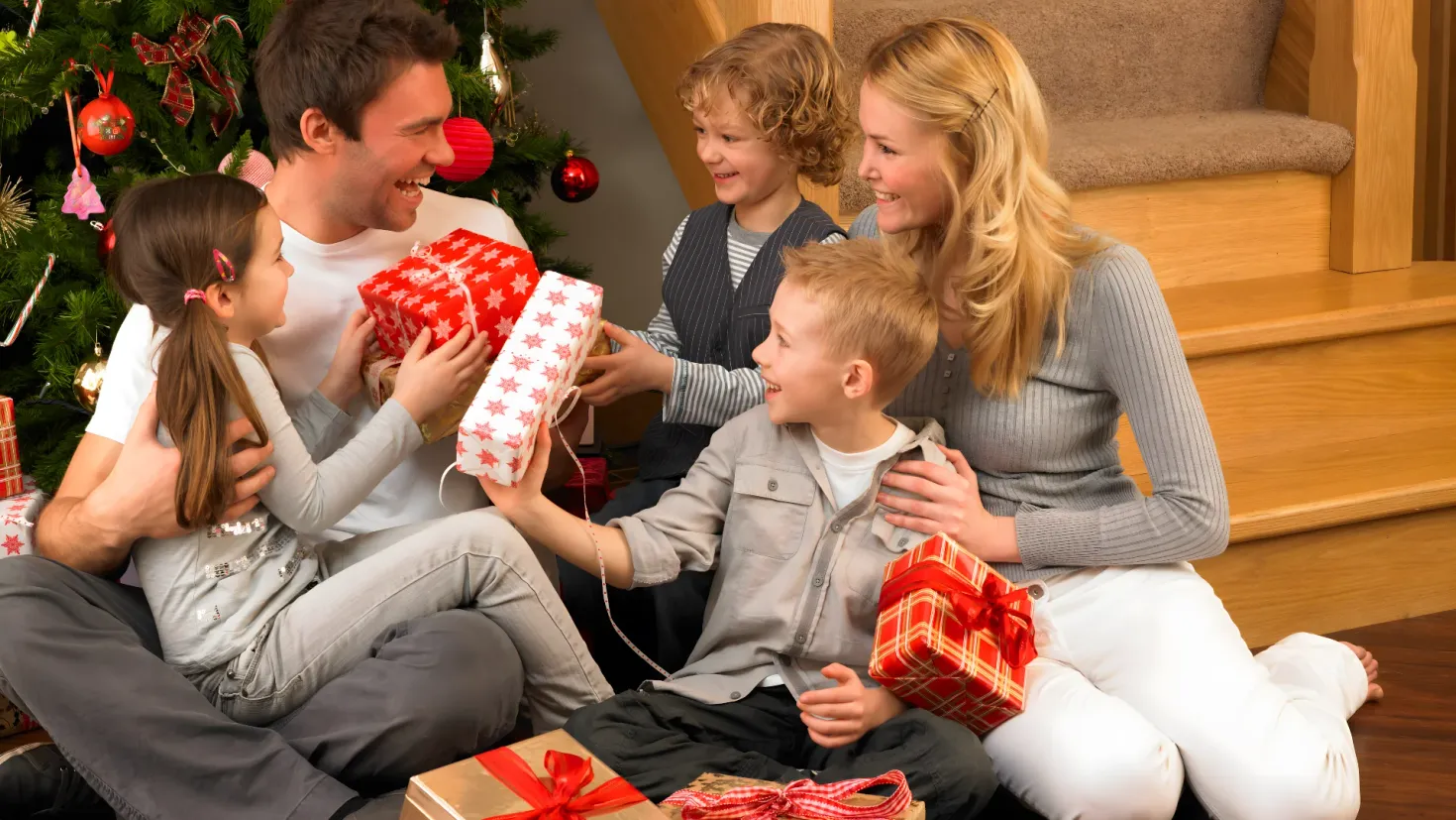 A family of four sitting on the floor in front of a Christmas tree, each holding a gift box in their hands. The tree is decorated with colorful ornaments and lights, and there is a staircase in the background.