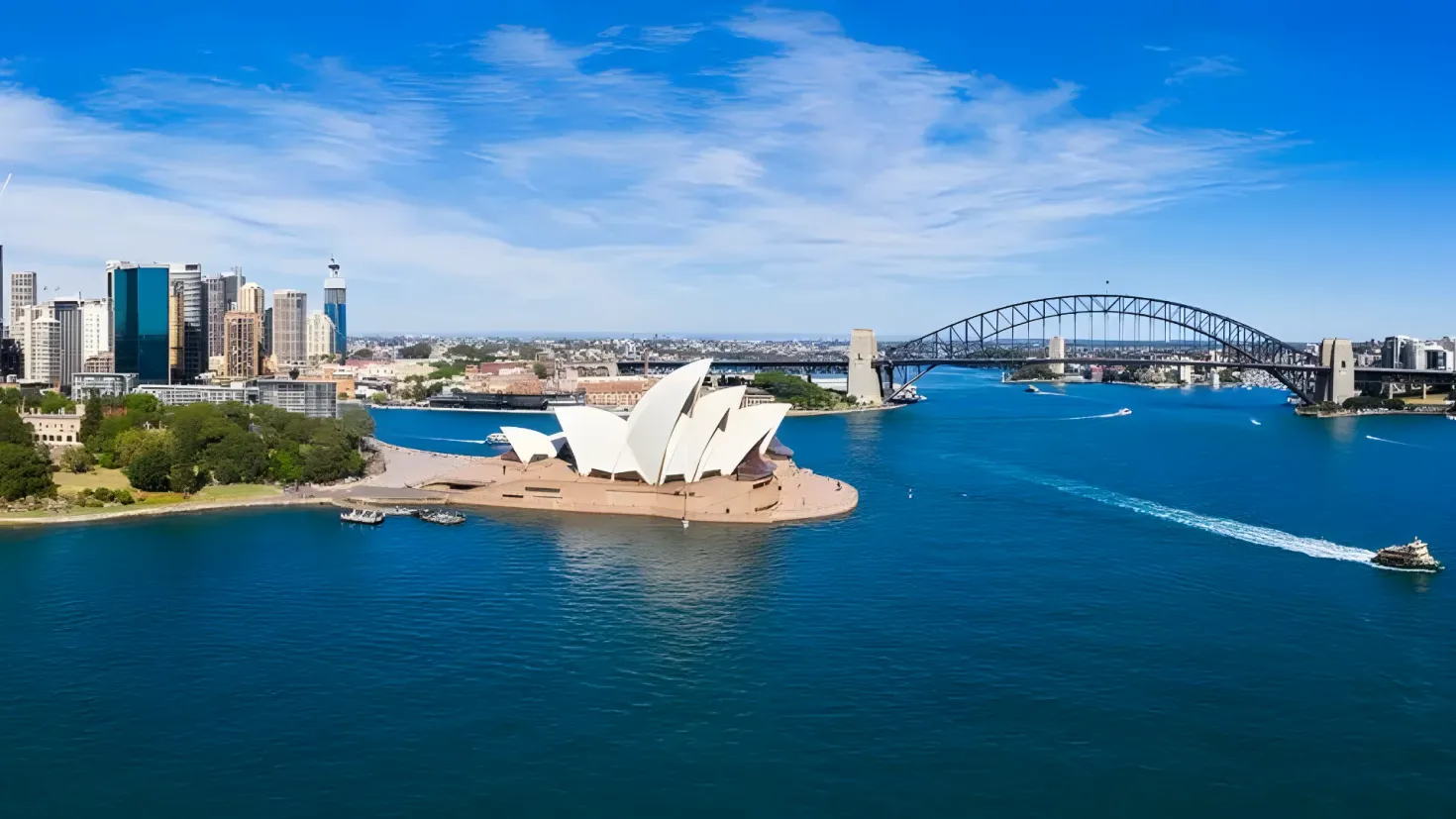 The Sydney Opera House and Harbour Bridge in Australia, with boats on the water, trees and buildings on the left side, and a sky with clouds in the background.