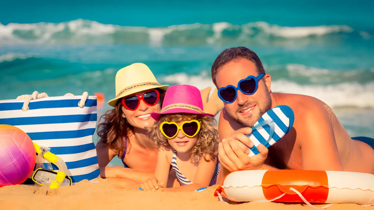 A happy family of three - a man, woman and two children - lying on the beach with their arms around each other. The man is holding an object in his hand, while the two children are wearing hats and goggles. On the left side of the image, there is a bag and a ball, and on the right side, there are tubes. In the background, the sea and sky can be seen.
