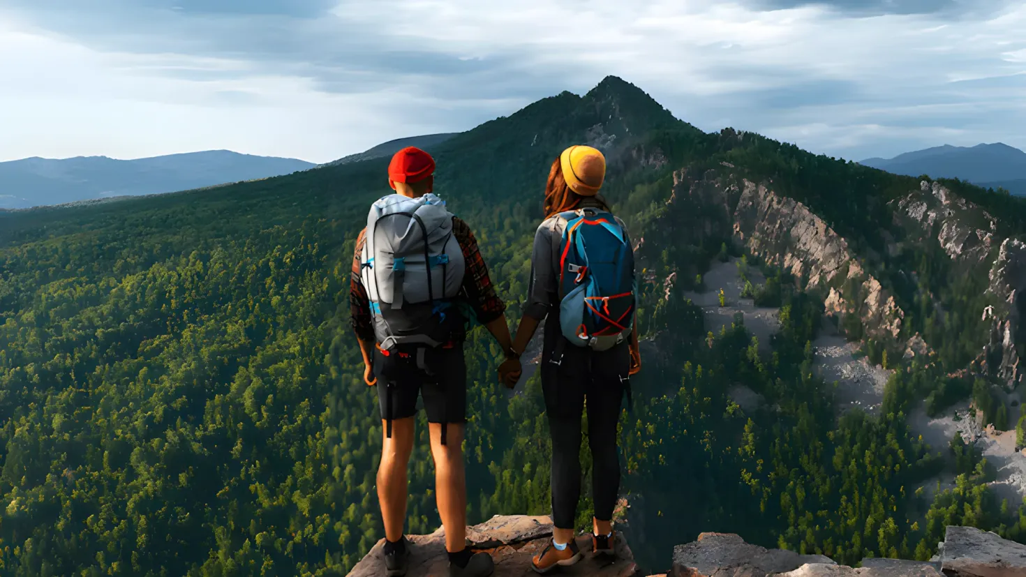 Two people standing on top of a mountain, holding hands and looking out at the view. They are both wearing backpacks and one of them is wearing a cap. The backdrop of the image is filled with trees, hills, and a sky with clouds.