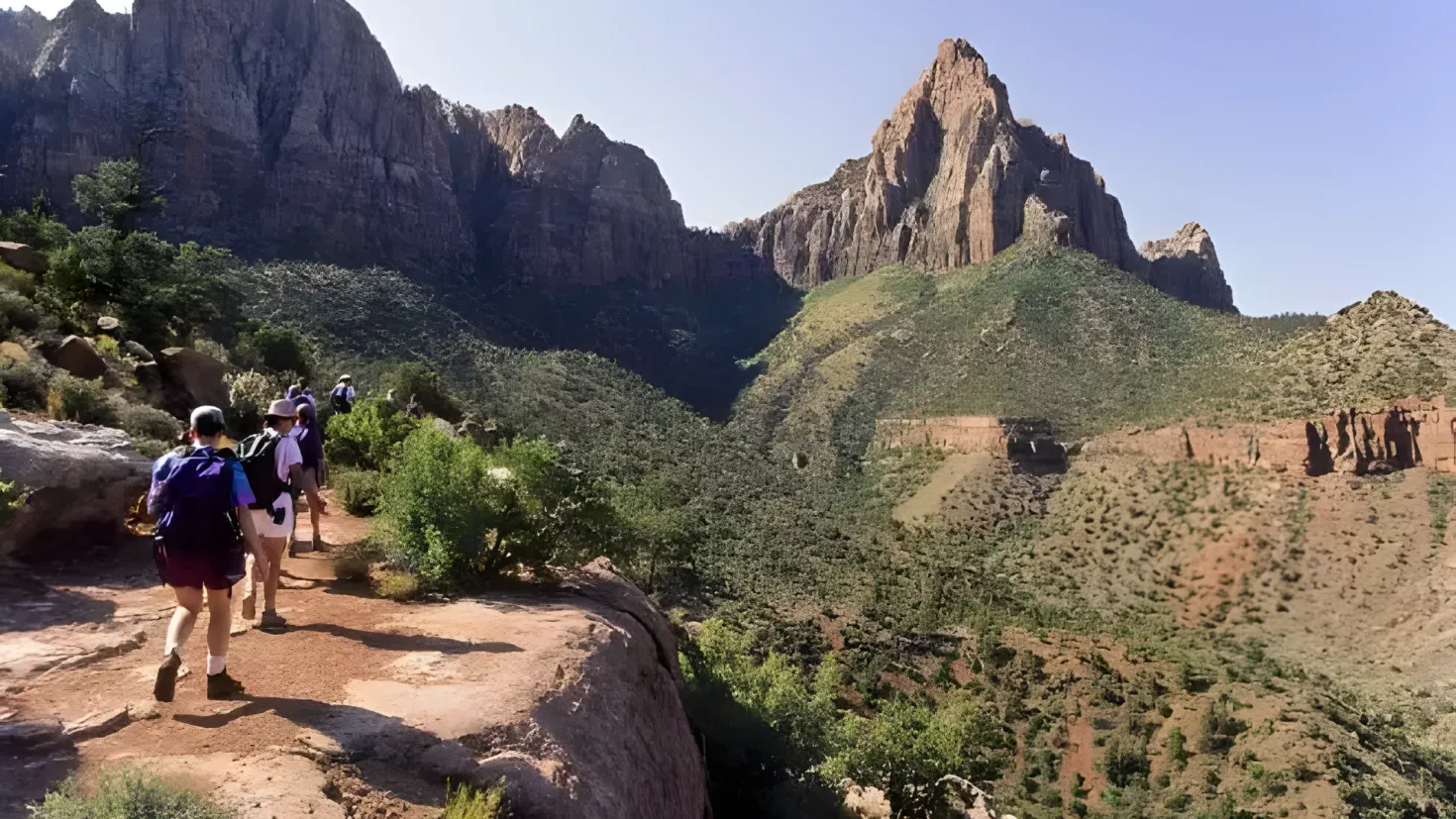 A group of people hiking up a rocky trail in Zion National Park, Utah. They are wearing backpacks and are surrounded by trees and hills. The sky is visible in the background.