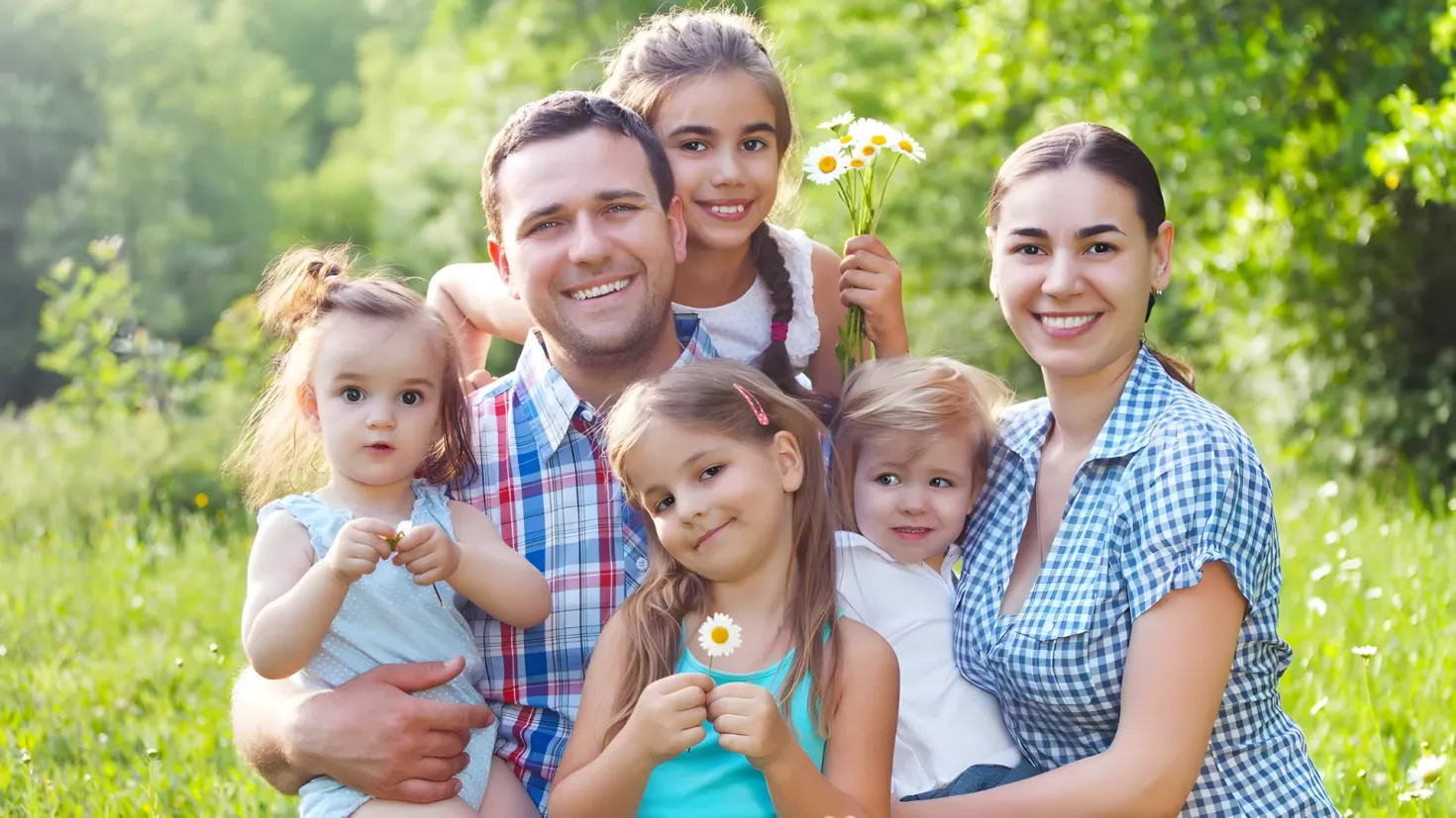 A happy family of five posing for a photo in a field of flowers. The man and woman are standing and smiling, while the two girls are holding daisies in their hands. The background of the image is filled with lush green plants and trees, creating a peaceful atmosphere.