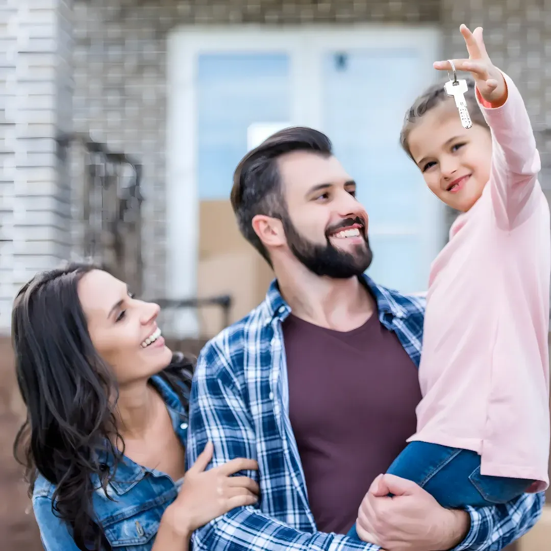 A happy family of three - a man, woman and a girl - standing in front of their new home, smiling and holding a key in their hand. The background is slightly blurred, suggesting that the focus of the image is on the family and the key they are holding.
