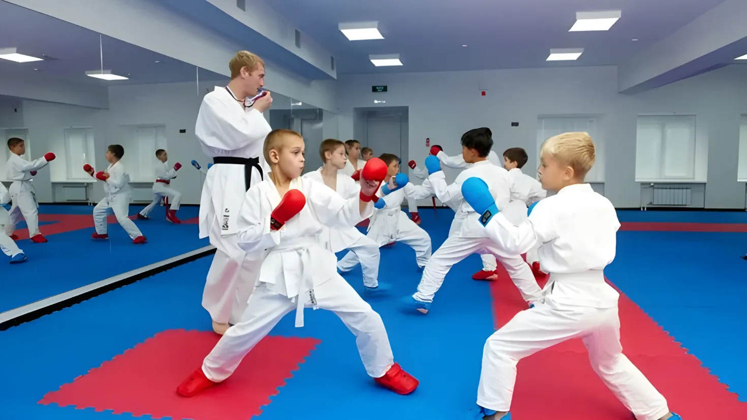 A group of children in karate gear practicing karate in a gym. We can see a man standing in the center of the group, surrounded by the children on the floor. The walls are adorned with windows and doors, and the ceiling is illuminated with lights.
