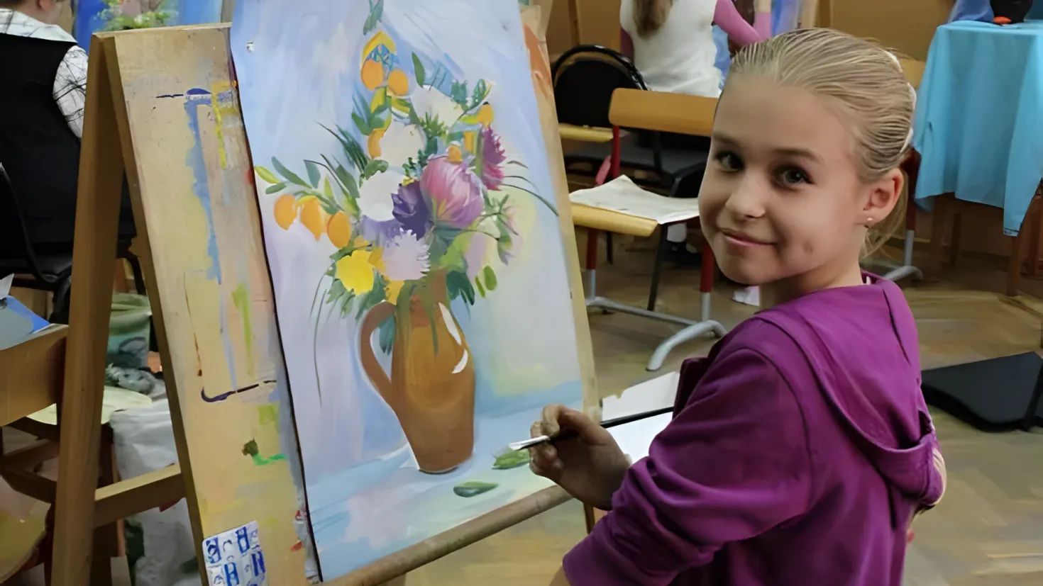 A young girl painting on an easel in a classroom. She is holding a brush in her hand and is surrounded by a group of people sitting on chairs around the easel. On the right side of the image, there is a table covered with a blue cloth and various objects on it. In the background, there are cardboard boxes and a wall.