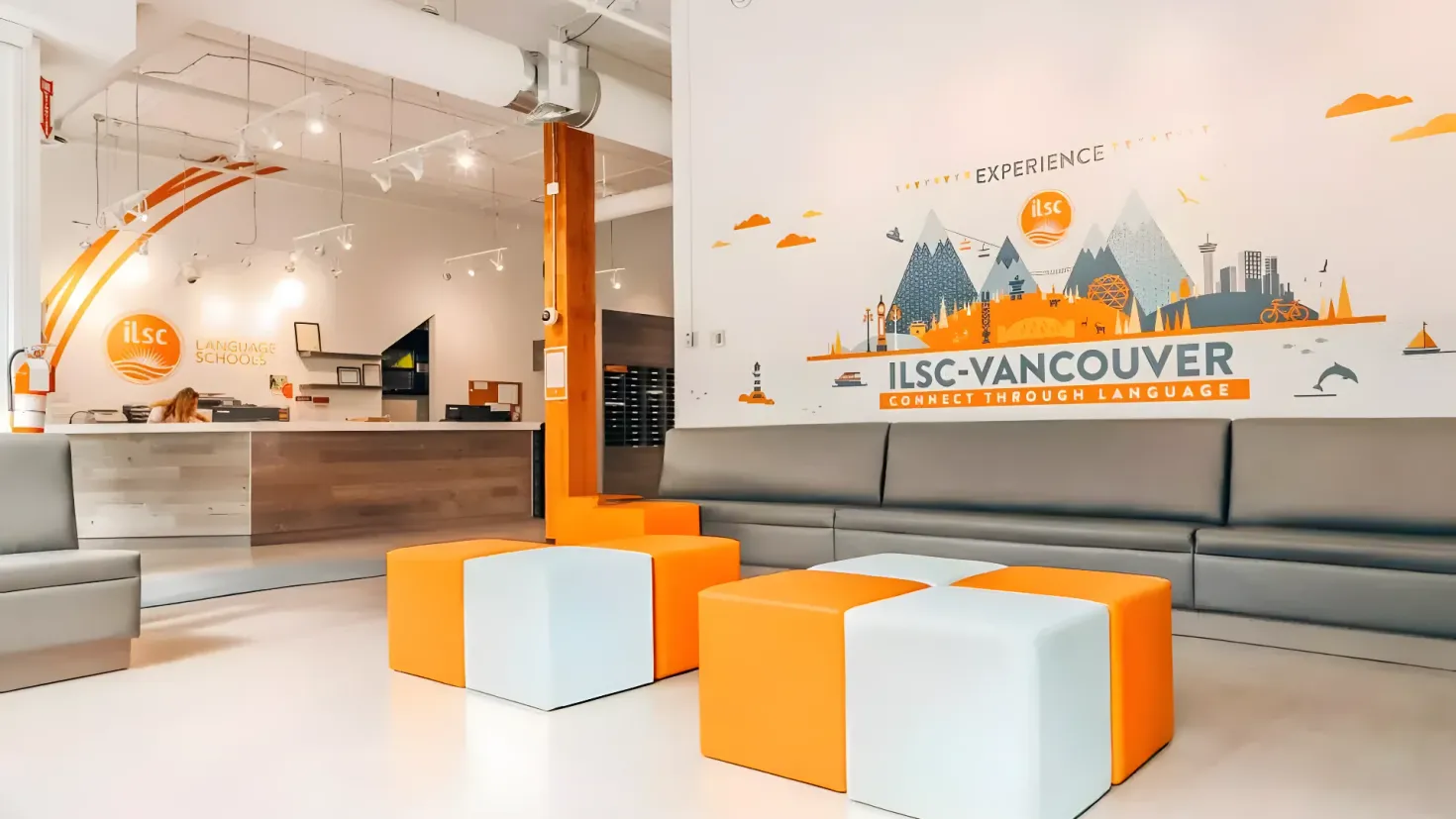 A reception area with orange and white furniture, including a studio couch, stools, and a desk. There is a person sitting on the couch, and in the background there is a wall with something written on it. The ceiling is adorned with lights, giving the room a warm and inviting atmosphere.