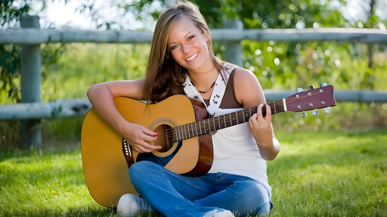 A woman sitting on the grass playing an acoustic guitar with a smile on her face, surrounded by a wooden fence and trees in the background.