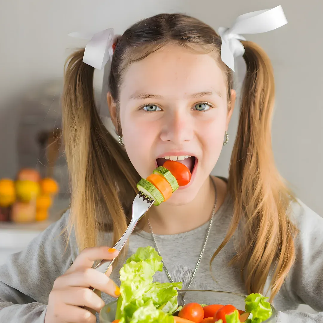 A young girl sitting at a table with a plate of vegetables in front of her. She is holding a fork in her hand and has a look of concentration on her face as she eats the vegetables. The background is slightly blurred, suggesting that the focus of the image is on the girl and her meal.
