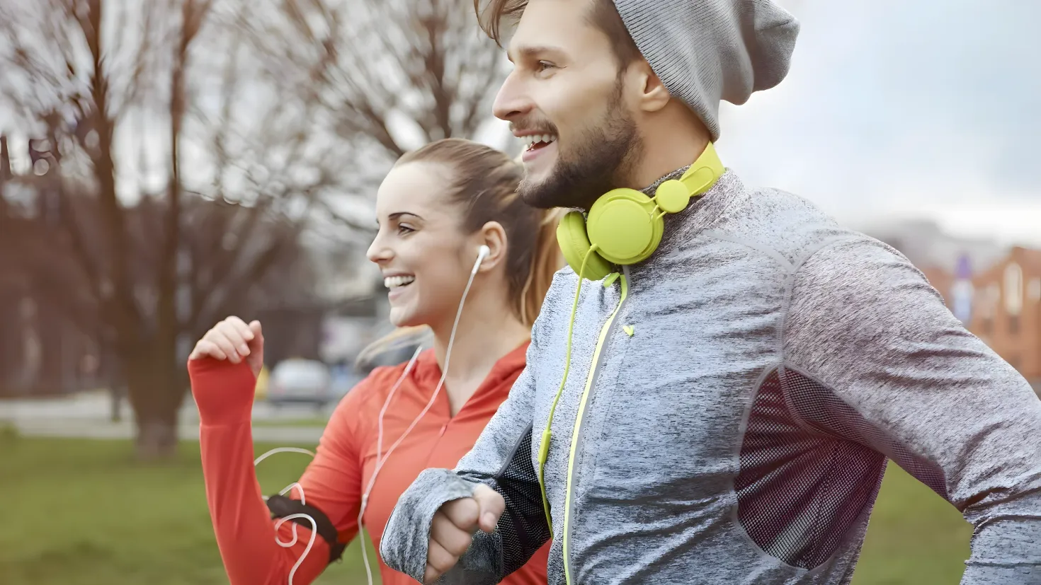 A man and woman jogging in a park, both wearing headphones and smiling. The background is slightly blurred, with trees, buildings, and a clear blue sky visible.