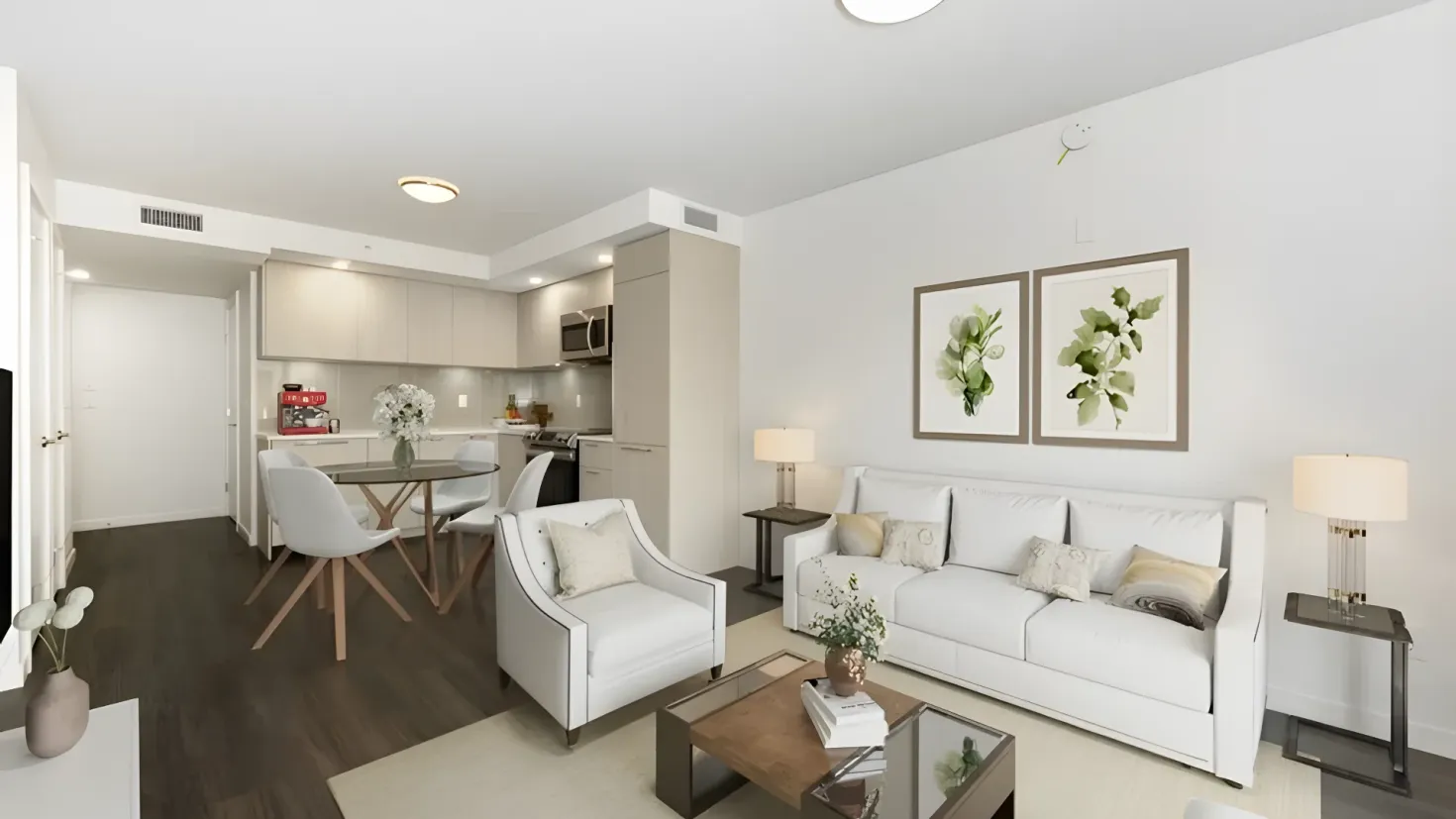 A living room filled with furniture such as a sofa set with cushions, side tables with table lamps, flower vases, a carpet on the floor, wall hangings attached to the walls, electric lights to the roof, a dining table with chairs, a flower vase on the table, a microwave oven, a gas stove, a dishwasher, a refrigerator, cupboards, and a door.