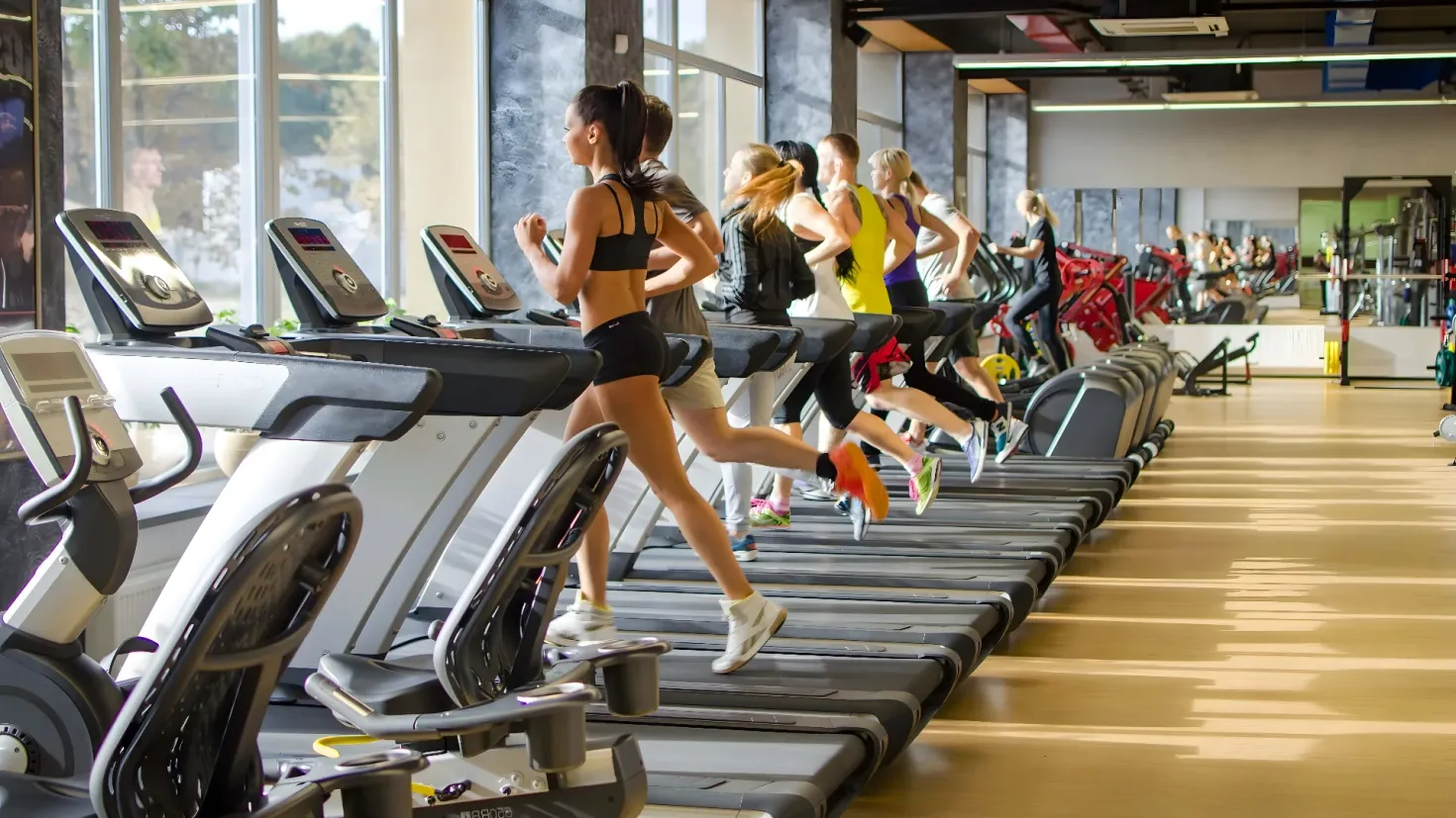 A group of people running on treadmills in a gym. There is a mirror on the left side of the image, reflecting the trees and sky outside. On the right side, there are a few people standing, and the ceiling is visible at the top of the picture.