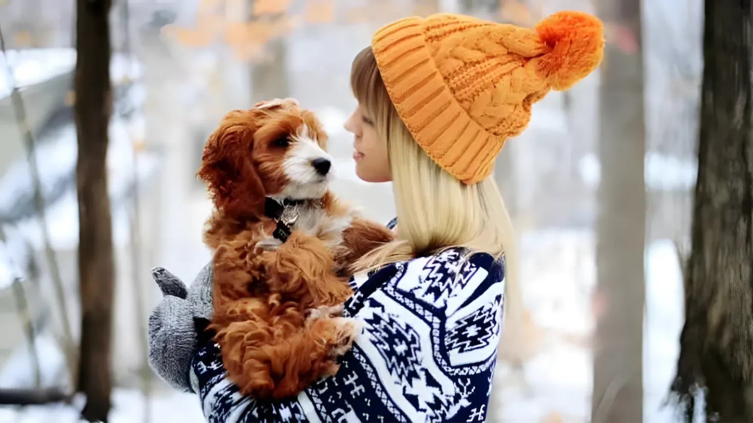 A woman wearing a blue and white dress and an orange cap, holding a brown and white dog in her arms, with a few trees in the background covered in snow.