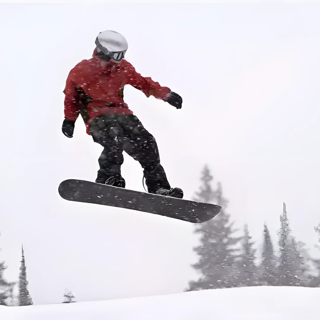 A snowboarder in mid air, wearing a red jacket, black pants, a white helmet, and black gloves. The snowboard is black and the background is filled with trees and a clear blue sky.