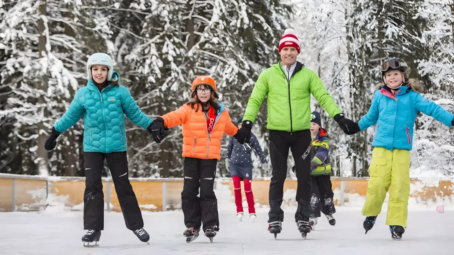 A family of four skating on an ice rink in the snow. They are all wearing jackets, helmets, gloves, and skating shoes, and the background is filled with trees covered in a blanket of snow.