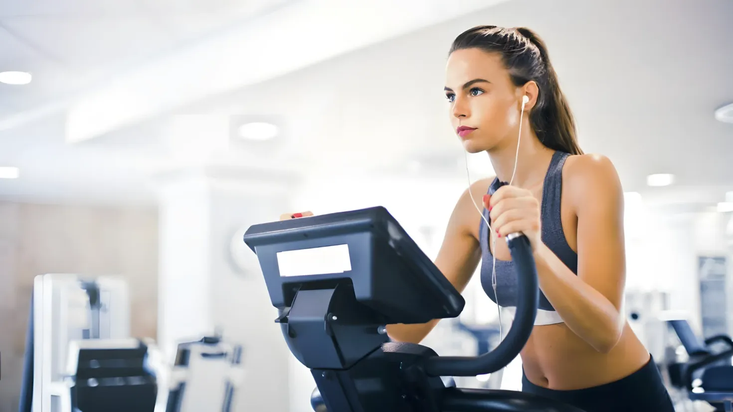 A woman on a stationary bike in a gym, wearing earphones and surrounded by other gym equipment. The background is slightly blurred, and the ceiling is visible at the top of the image.