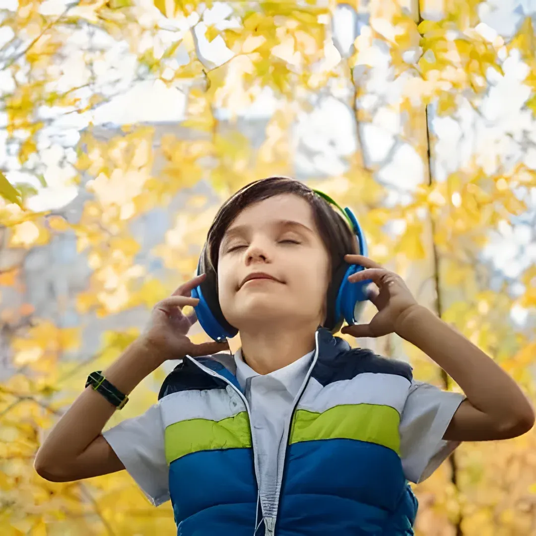 A young boy wearing headphones and listening to music in the park. He is standing in front of a backdrop of trees and a clear blue sky.