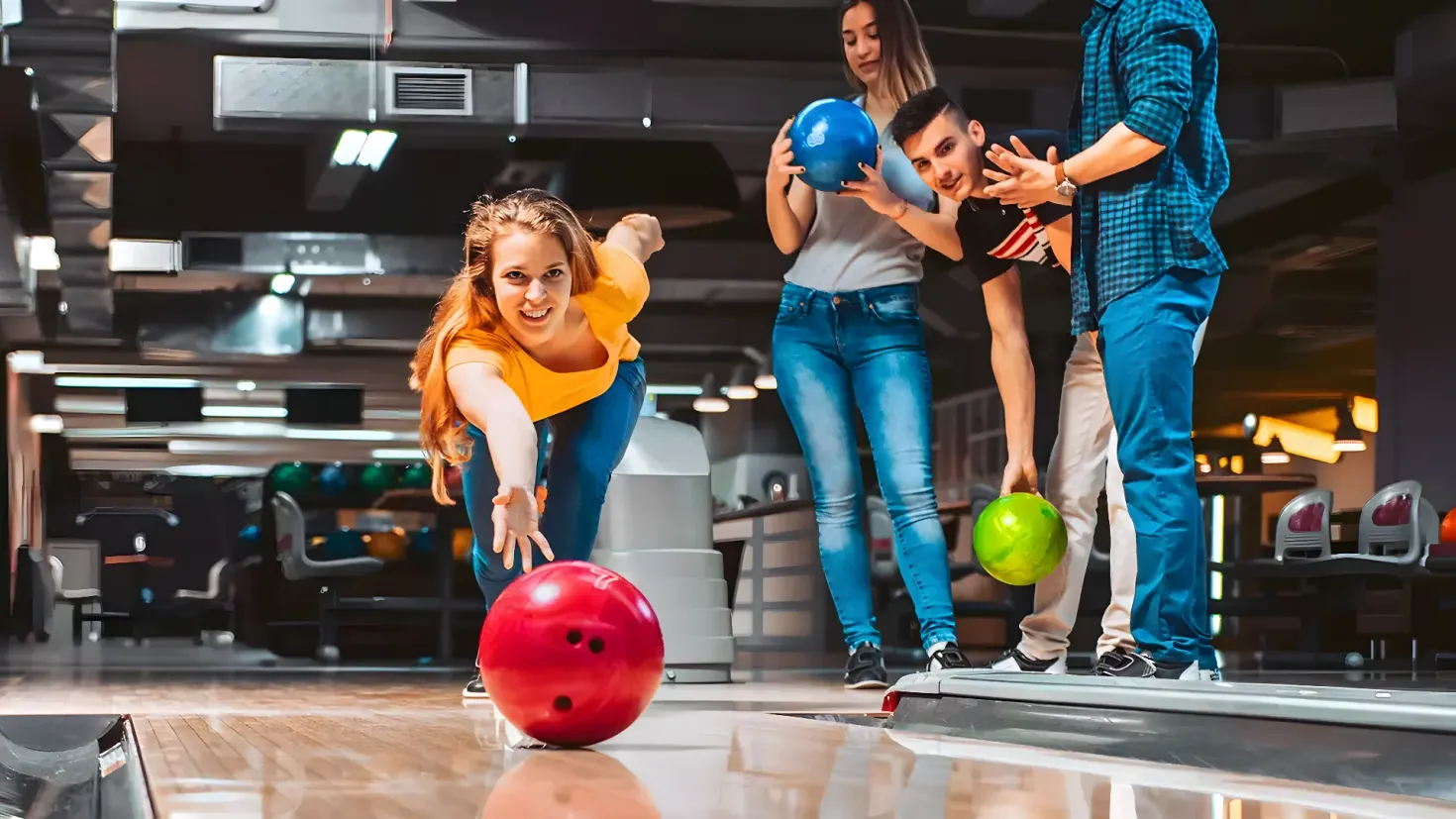 A group of four people playing bowling in a bowling alley. The woman in the center is wearing a yellow t-shirt and blue pants, and is holding a bowling ball in her hand. The other three people are standing around her, each holding a different colored bowling ball. In the background, there are several bowling balls, chairs, and lights on the ceiling.