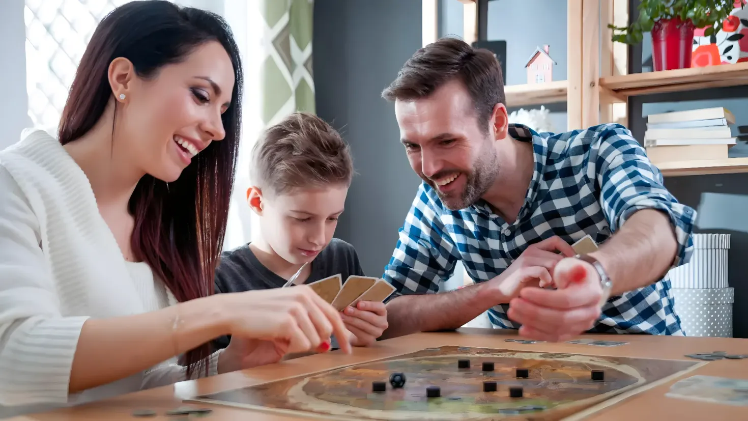 A family of three - a man, woman and a boy - playing a board game together. On the table in front of them are coins and cards, and to the right of them is a chair. In the background, there is a cupboard with books, a flower vase and other objects, as well as a window with a curtain.