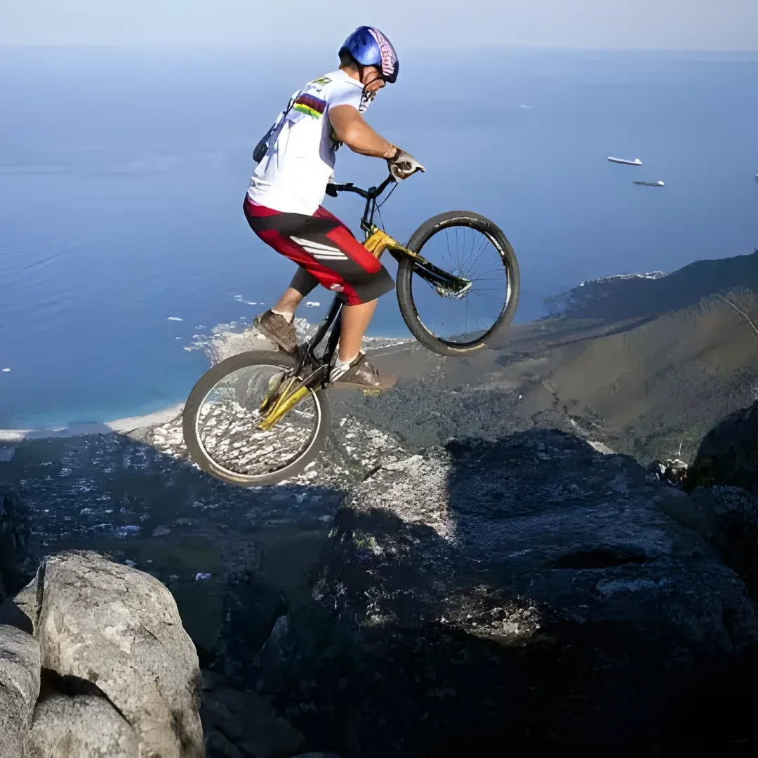 A man riding a mountain bike on top of a rocky cliff, wearing a white t-shirt, red shorts, and a blue helmet. In the background, there is a body of water with boats sailing on it, and the sky is a clear blue.