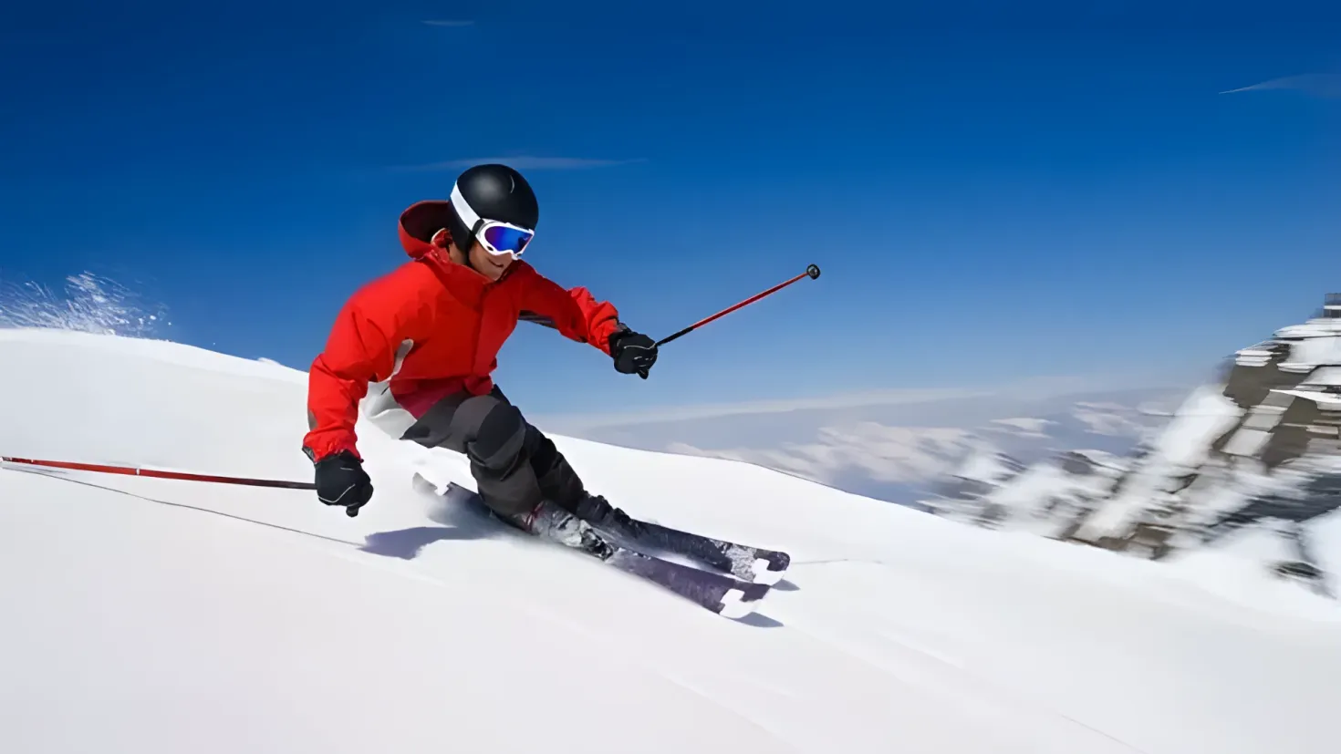A person in a red jacket skiing down a snowy hill, wearing a helmet, goggles, gloves, and shoes, and holding ski poles. The sky is visible in the background.