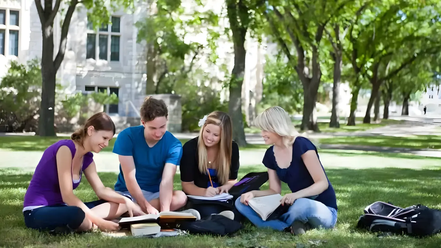 A group of four young people sitting on the grass in front of a building, surrounded by trees and a clear blue sky. They are all holding books in their hands and there are bags scattered around them.