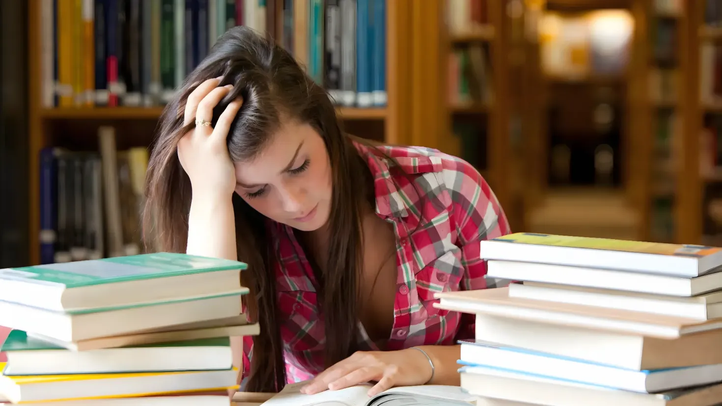 A young woman sitting at a table in a library, surrounded by bookshelves filled with books. She appears to be deep in thought, perhaps contemplating the five reasons why she is a student.