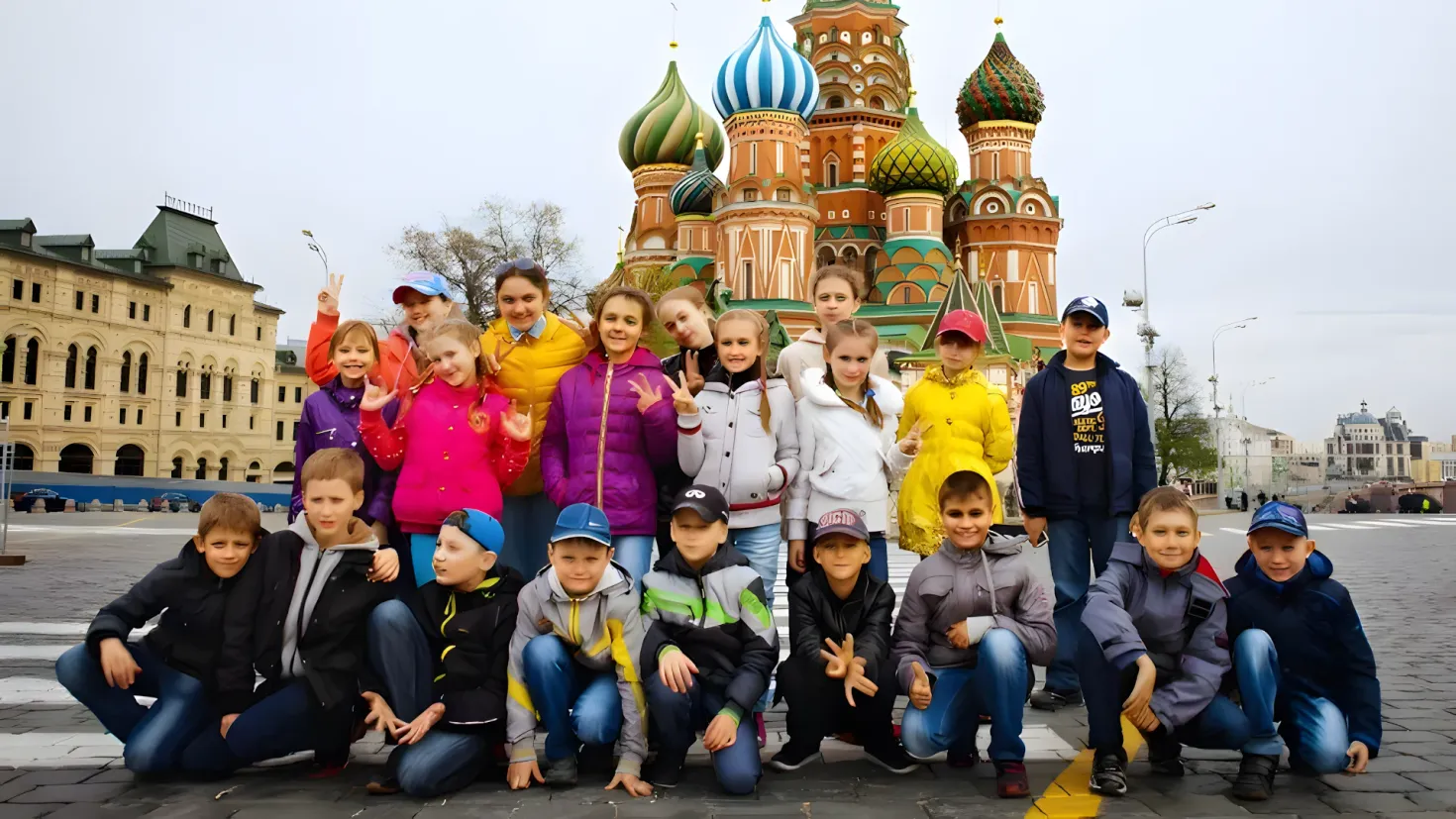 A group of children posing for a photo in front of St. Basil's Cathedral in Moscow, Russia. Some of the children are wearing caps, while others are in a squatting position. In the background, there are light poles, trees, buildings with windows, and a clear blue sky.