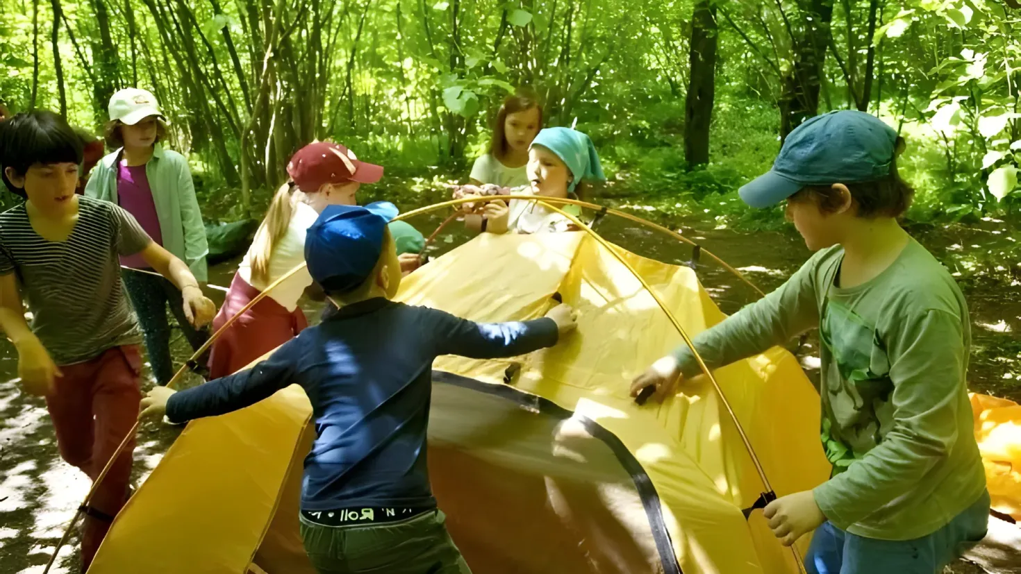 A group of children standing around a yellow tent in the woods. Some of the children are wearing caps, and there are trees in the background.