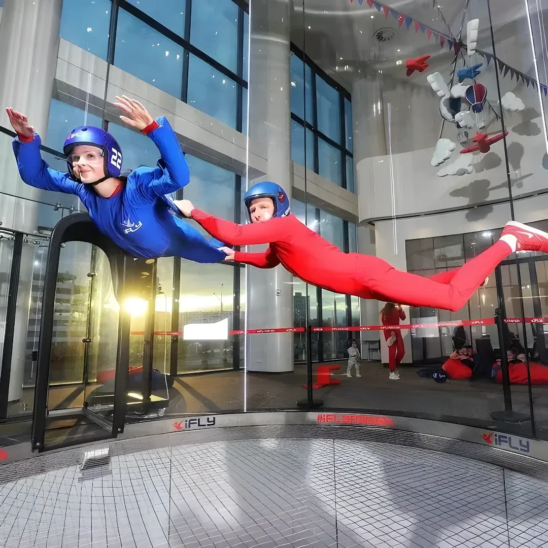Two people in blue and red skydiving in a large building with glass walls and pillars. They are both wearing helmets and sneakers, and there are a few other people in the background.