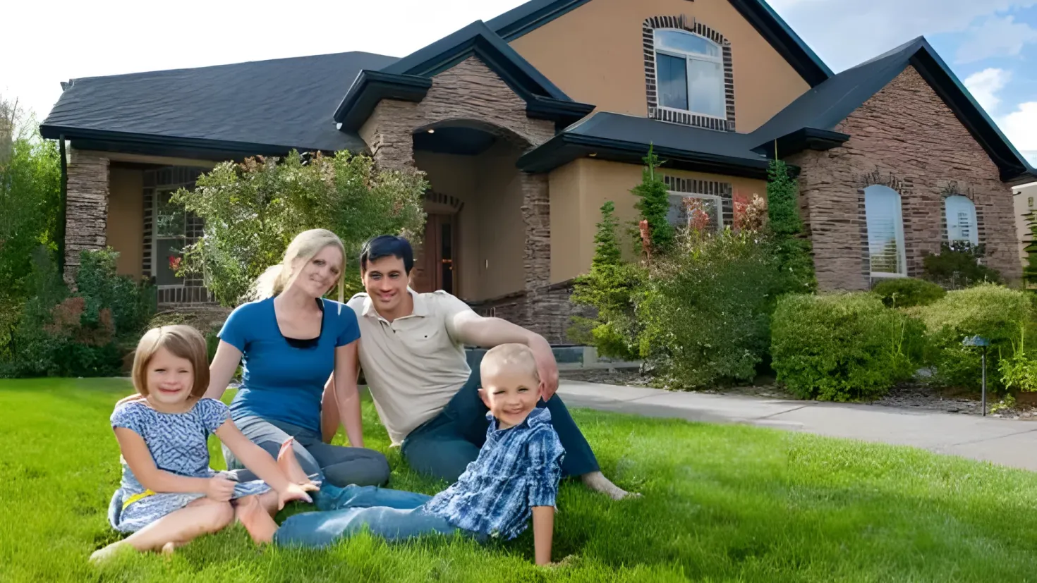 A family of four - a man, woman and two children - sitting on the grass in front of a house. The house is surrounded by trees and plants, and the sky is visible in the background.
