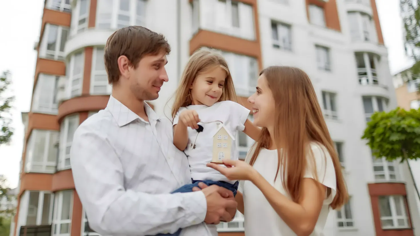 A happy family of three - a man, woman and a little girl - standing in front of a building with windows, surrounded by trees and a clear blue sky. The man is holding a toy house in his hands, suggesting they are in the process of buying a new home.