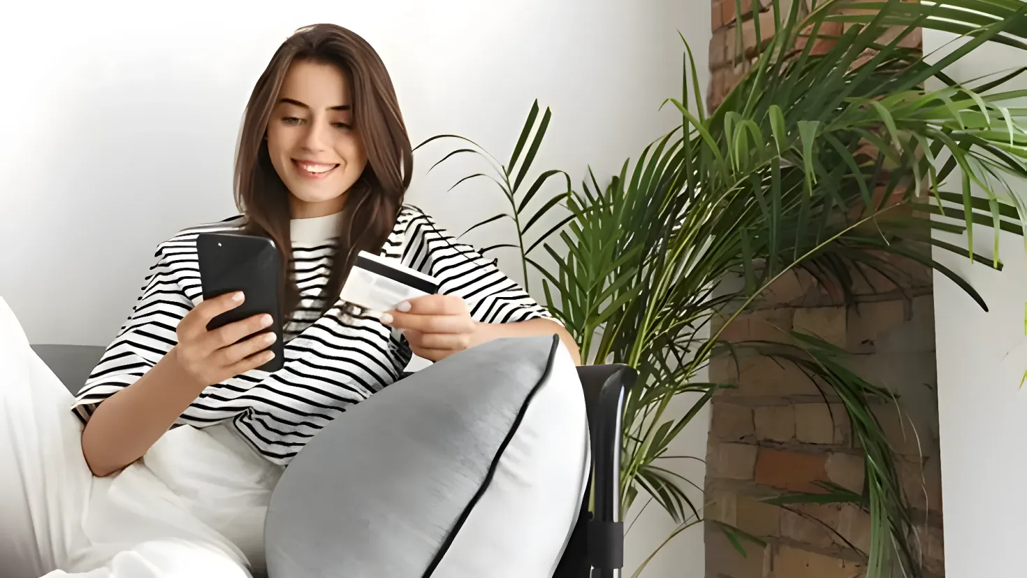 A woman sitting on a couch, holding a credit card in one hand and a mobile phone in the other. She is surrounded by a pillow and a plant, and there is a wall in the background.