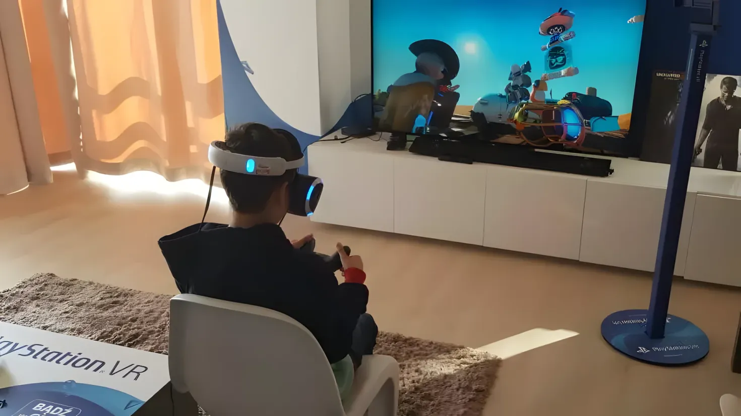 A person sitting in a chair in front of a large screen, wearing a virtual reality headset. On the right side of the image, there is a television on a stand, and on the left side, there are curtains. The floor is covered with a carpet.