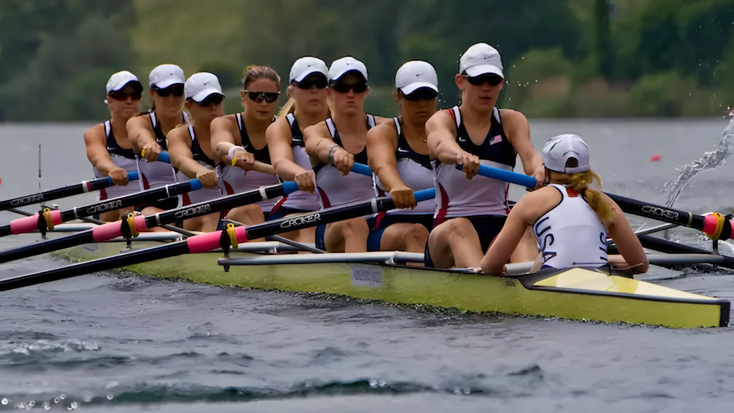 A group of women rowing on top of a body of water, wearing caps and goggles, and holding paddles in their hands. In the background, there are trees providing a scenic backdrop.