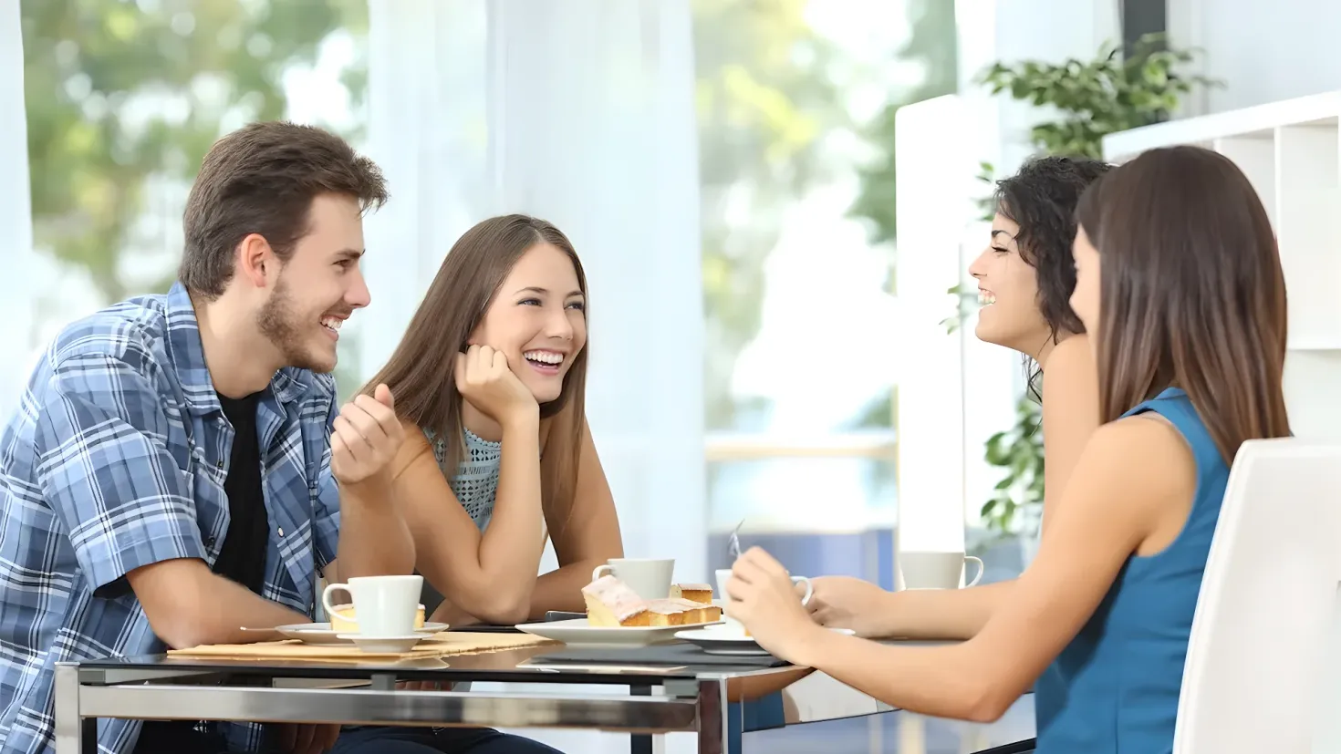 A group of four people sitting around a table in a restaurant, smiling and enjoying each other's company. On the table there are cups, saucers, plates, food items, and other objects. In the background, there is a plant and a cupboard, and the background is slightly blurred.