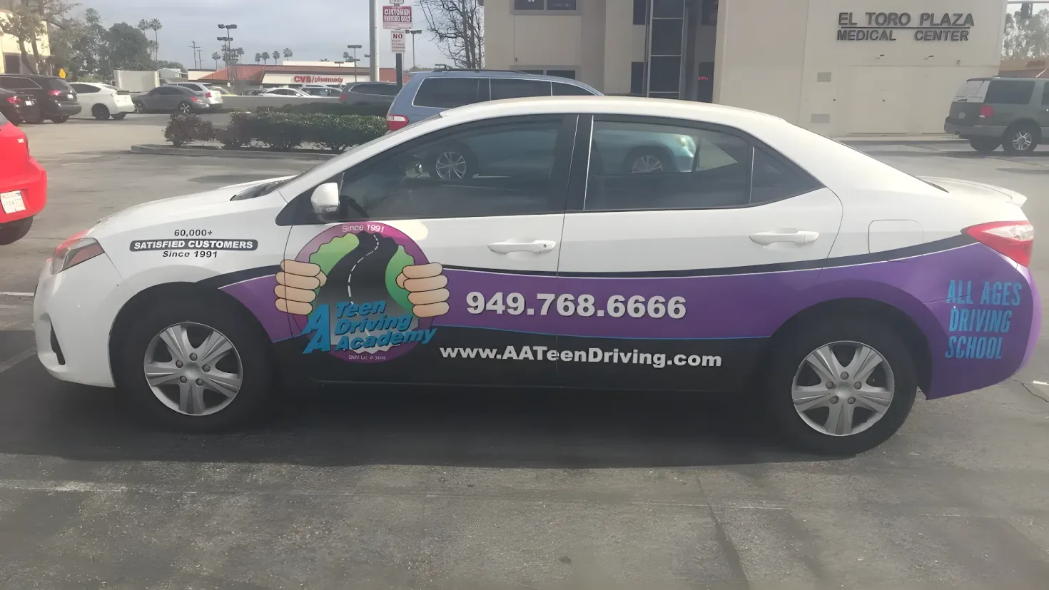 A car parked in front of a building with a sign on it. The car is surrounded by street light poles, trees, and plants, and the sky is visible in the background.