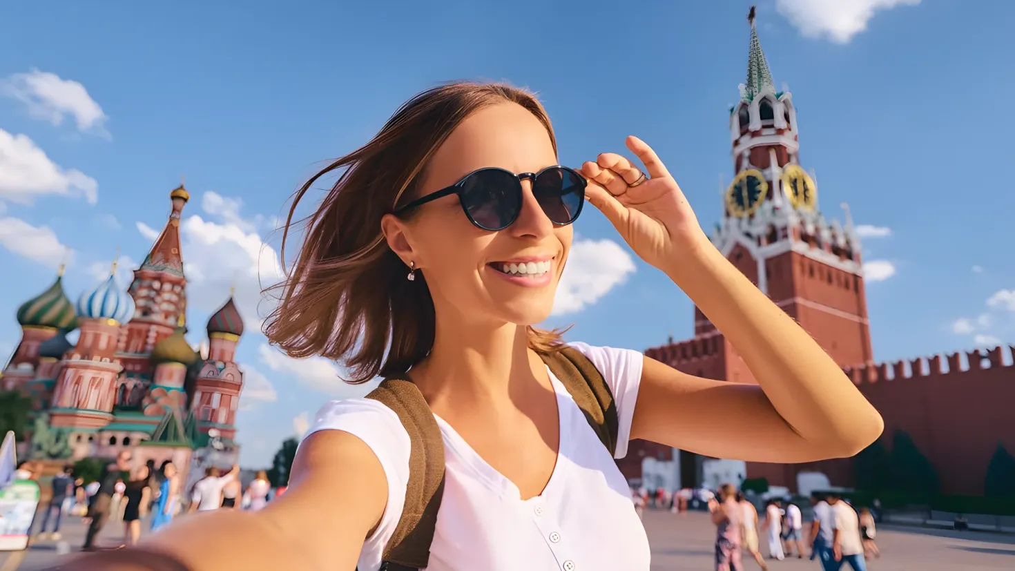 A woman taking a selfie in front of St. Basil's Cathedral in Moscow, Russia. She is wearing a white t-shirt and carrying a bag, and is smiling. In the background, there are a number of people walking on the road, trees, and the sky. On the left side of the image, there is a board with something written on it.