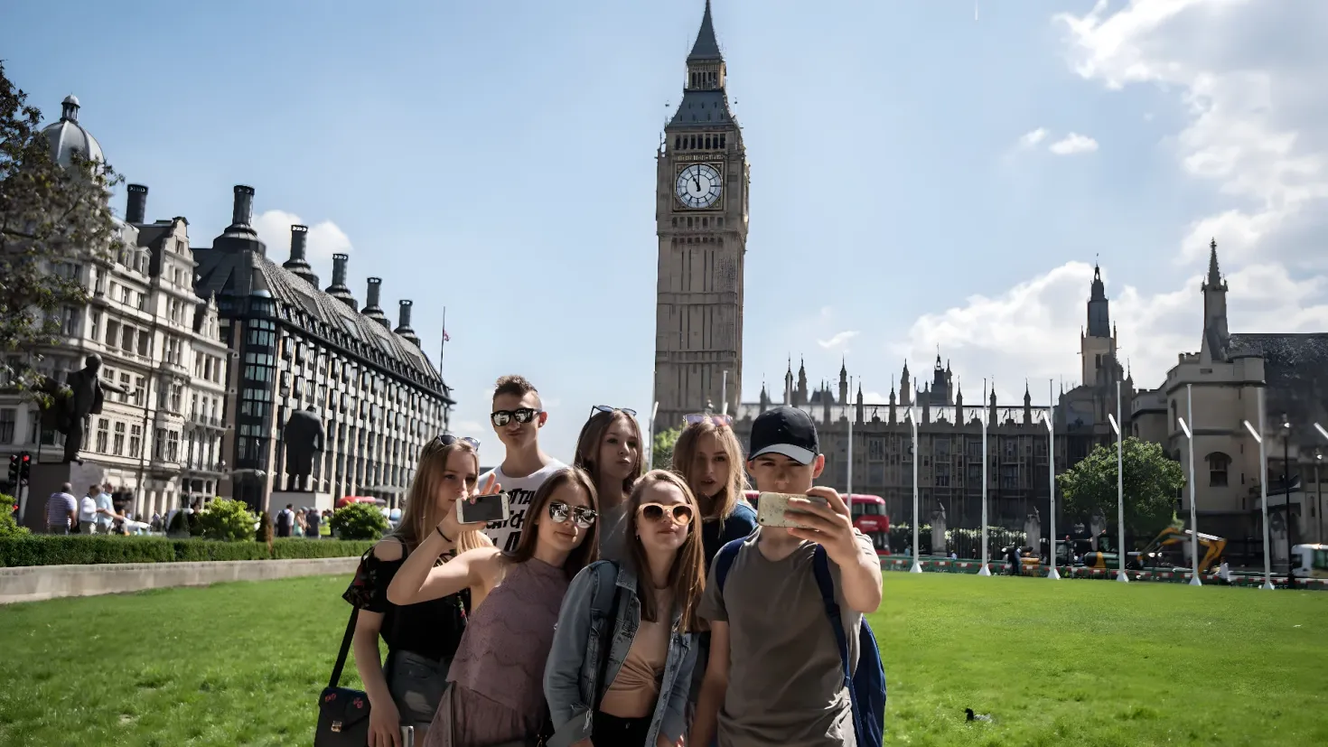 A group of people standing in front of Big Ben in London. Some of them are wearing goggles and carrying handbags, while others are holding mobile phones. The ground is covered in grass and there are trees and buildings with windows in the background. The sky is filled with clouds and there is a clock tower in the distance.