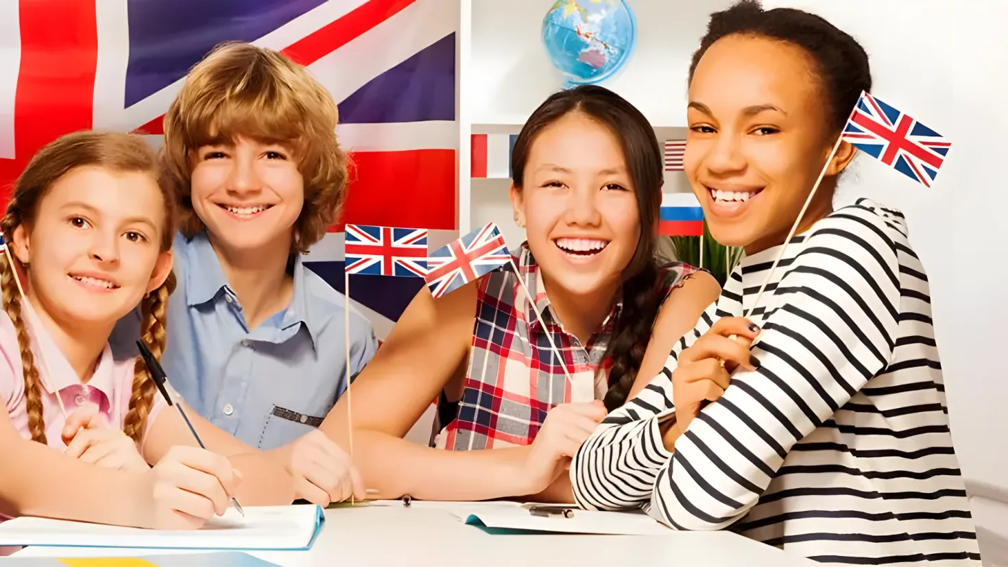 A group of four children sitting at a table with British flags in the background. On the table there are books and papers, and on the wall behind them there is a globe and flags. The children are smiling, suggesting that they are learning English.