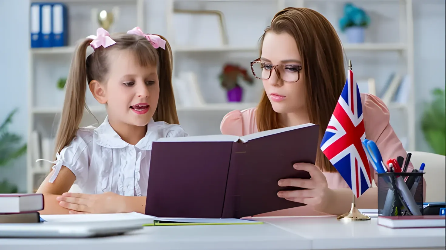 A woman and a little girl sitting at a desk, both wearing glasses. The woman is holding a book in her hands and the little girl is looking at it intently. On the desk there are books, pens in a pen stand, and other objects. In the background there are shelves with files and other items, as well as plants.
