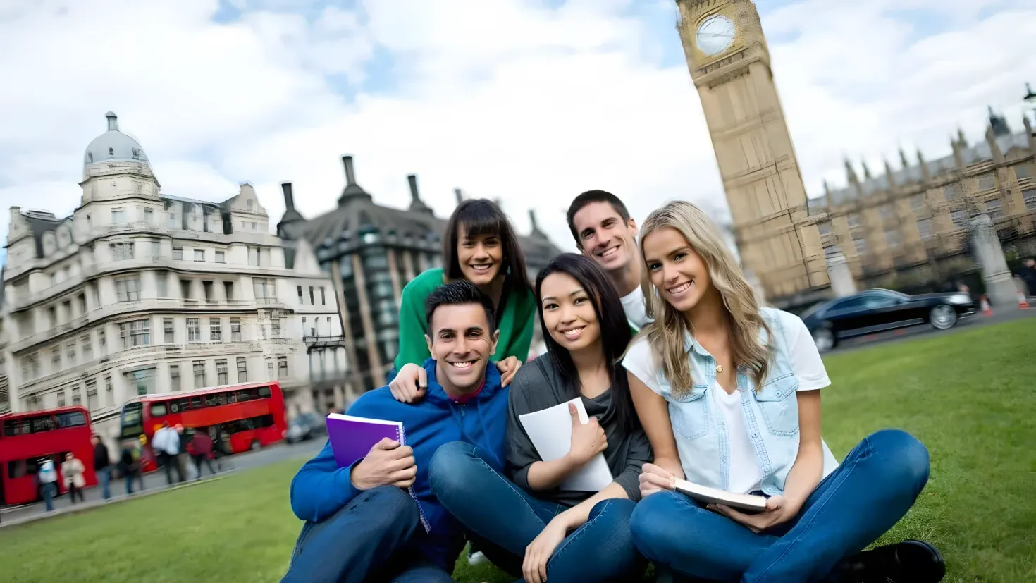 A group of young people sitting on the grass in front of Big Ben in London. They are all smiling and holding books in their hands. In the background, there are buildings, vehicles on the road, people walking on the footpath, traffic cones, and a clock tower. The sky is filled with clouds.