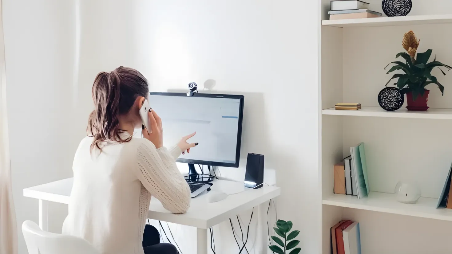 A woman sitting at a desk in front of a computer, holding a mobile phone in her hand. On the desk there is a monitor, keyboard, mouse, speakers, and other objects. To the right of the desk is a rack with books, a pot with a plant, and a few other items. In the background, there are curtains and a wall.