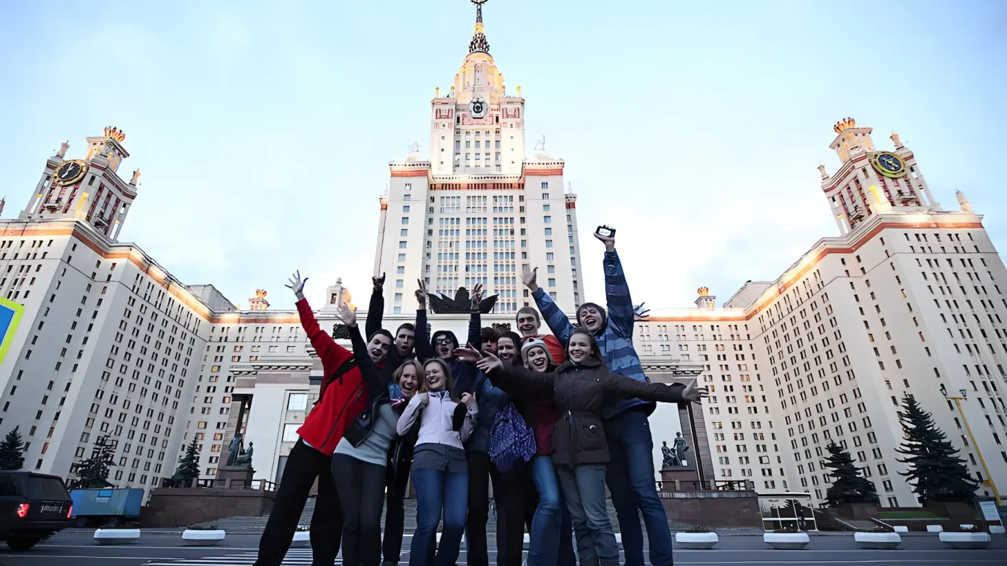 A group of people standing in front of a large building with a clock tower, surrounded by trees, light poles, statues, and a vehicle on the road. The sky is visible in the background.