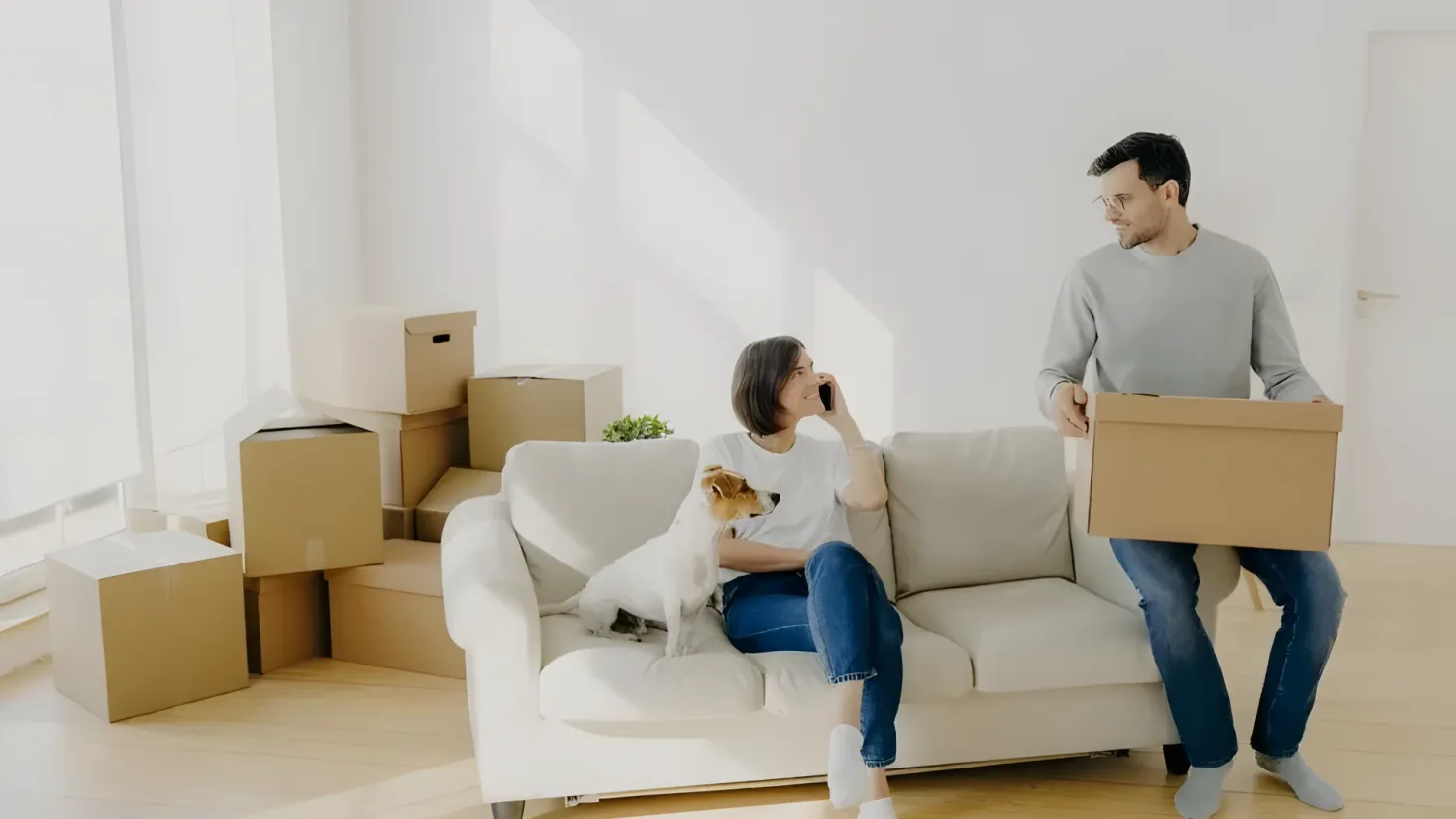 A man and woman sitting on a couch with a dog, surrounded by cardboard boxes. The man is holding a cardboard box in his hands, while the woman is holding an object in her hand. In the background, there is a wall and a door, suggesting they are in the process of moving into their new home.