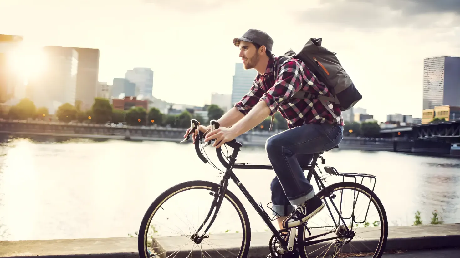 A man riding a bicycle down a sidewalk next to a river, with a backpack on his back. He is wearing a cap and the background of the image is filled with buildings, trees, and a bridge. The sky is visible at the top of the picture.