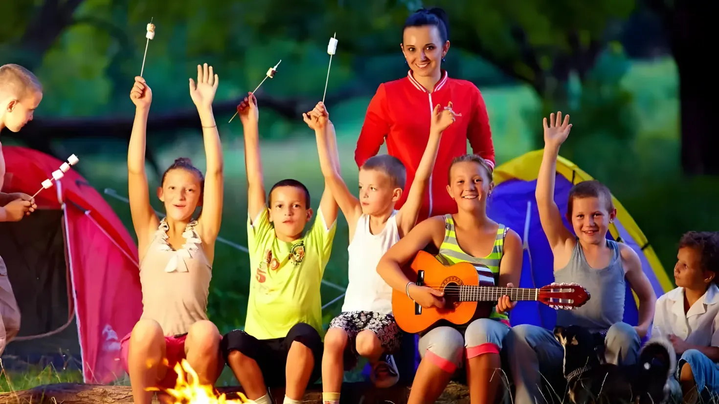A group of children sitting around a campfire with marshmallows in their hands, surrounded by grass and trees in the background. One of the children is playing a guitar, while the others are holding sticks.
