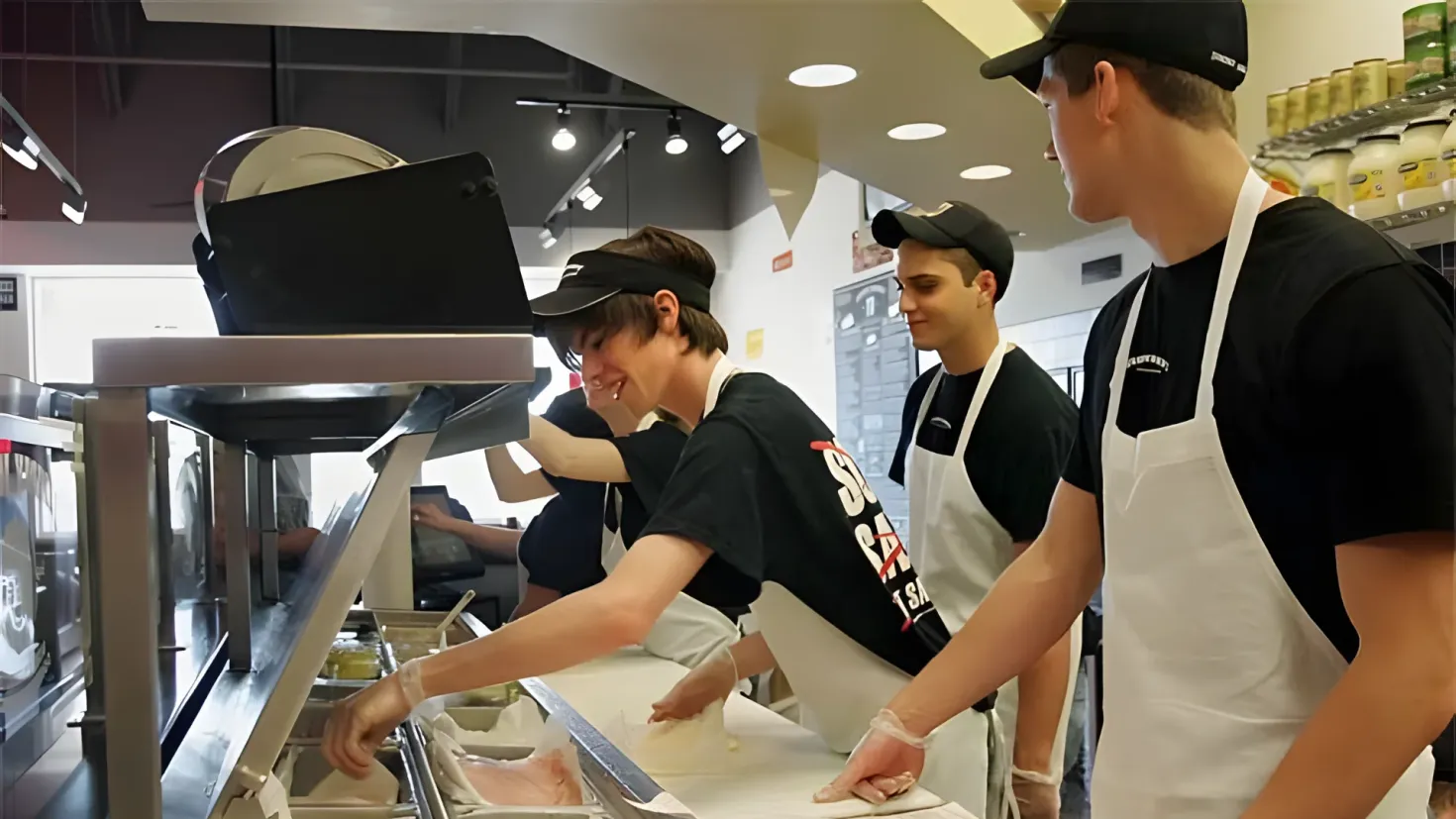 A group of people wearing caps and aprons standing around a counter in a kitchen. On the counter there are trays with food items, and on the right side of the image there is a rack with jars and other items. In the background there are lights on the ceiling.